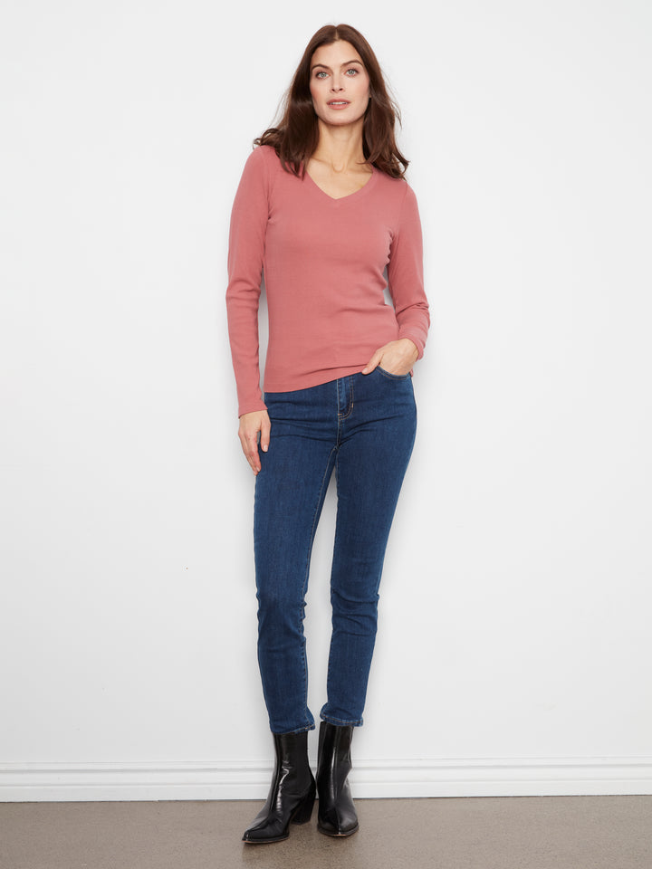 A woman modeling a dusty rose long-sleeve V-neck knit sweater paired with dark blue skinny jeans and black ankle boots, standing against a plain white wall.