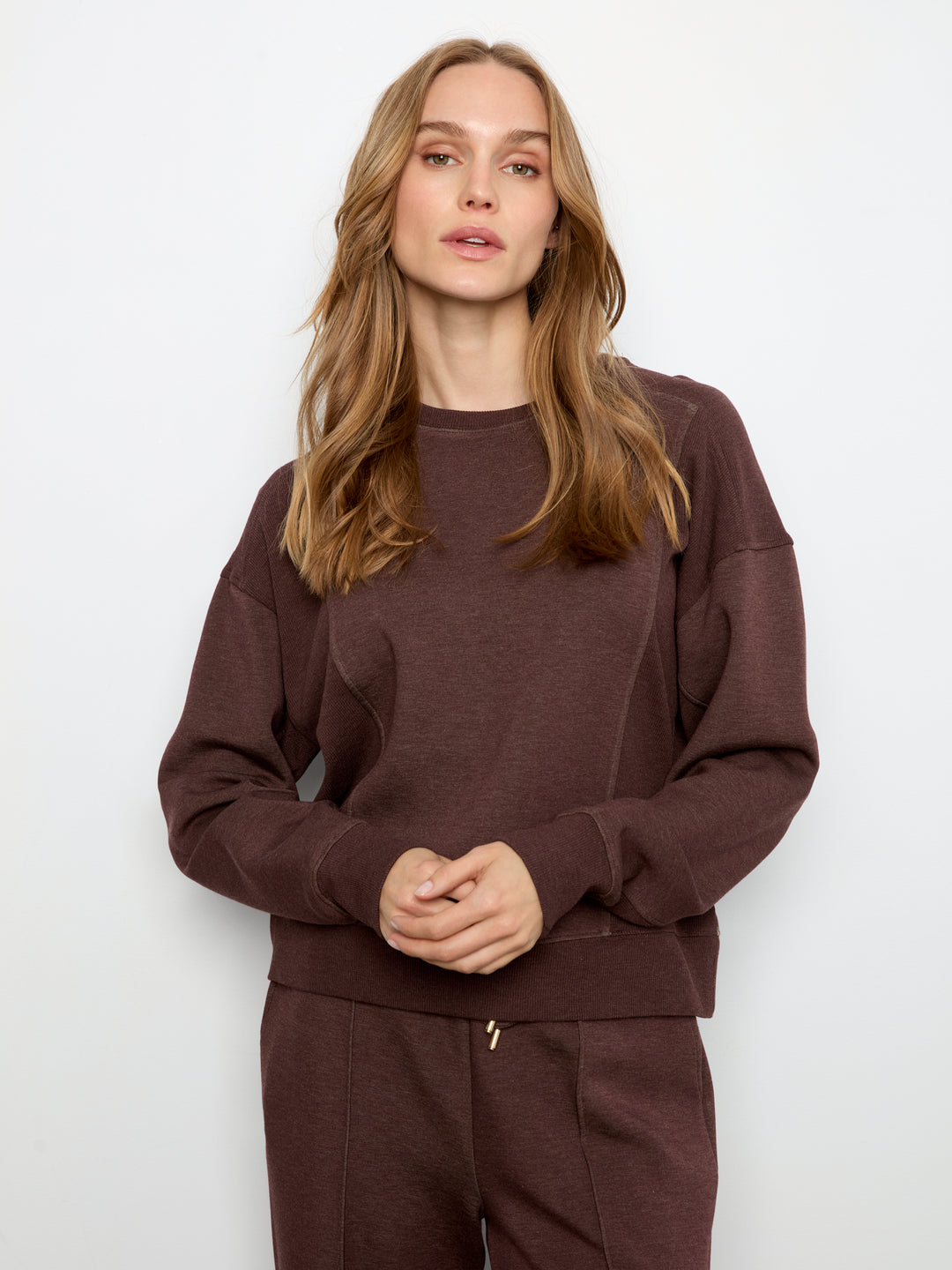 A woman modeling a dark brown crewneck sweatshirt and matching sweatpants against a plain white studio background.