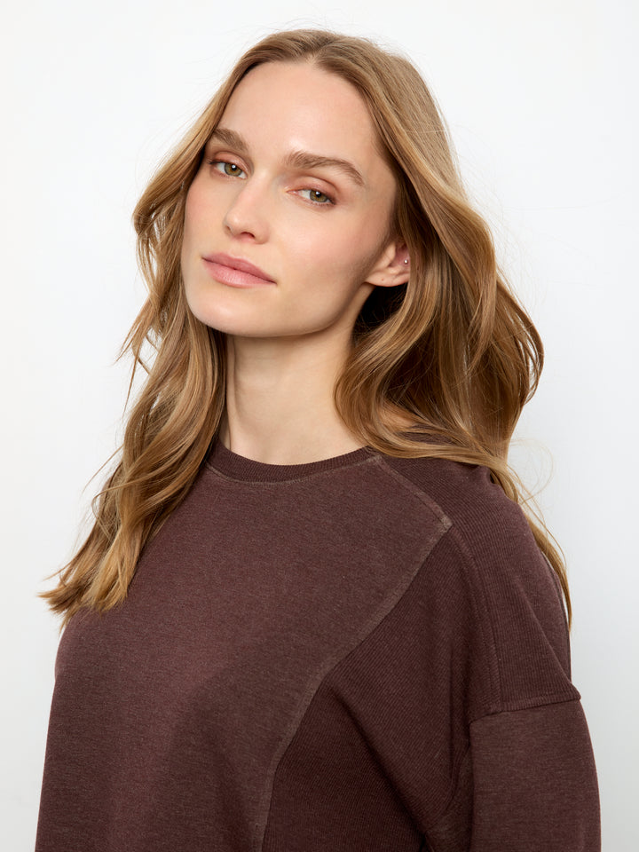 A woman with wavy light brown hair wearing a dark brown long-sleeve crewneck sweatshirt, photographed against a clean white background.