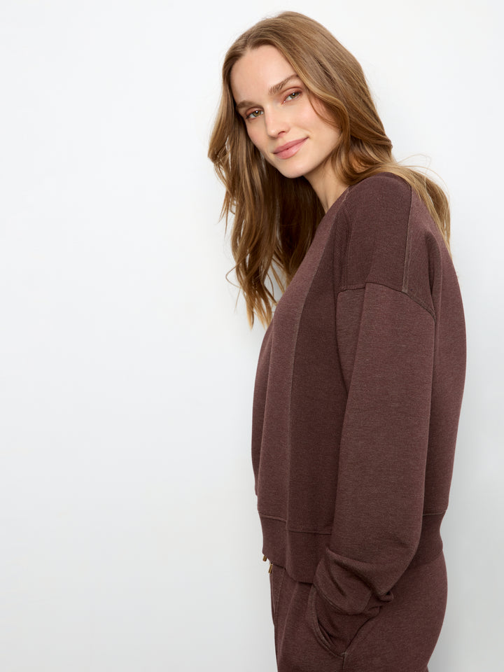 A woman wearing a dark brown long-sleeve knit top or sweatshirt, shown from the side against a plain white studio background.