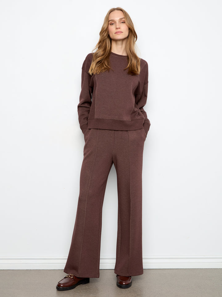 A woman modeling a matching brown knit sweater and wide-leg trousers set against a plain white wall.