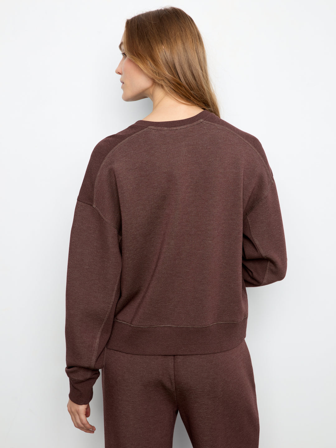 A woman shown from the back wearing a dark brown crewneck sweatshirt and matching sweatpants against a plain white studio background.