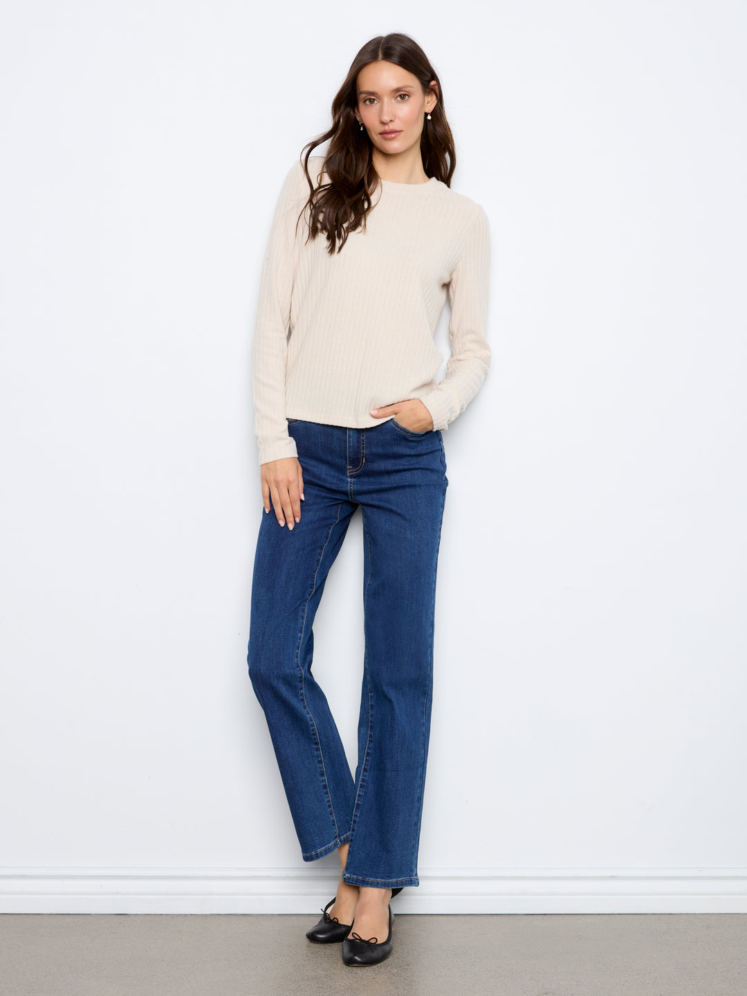 A woman modeling a cream long-sleeve knit sweater and dark blue straight-leg jeans against a plain white wall.
