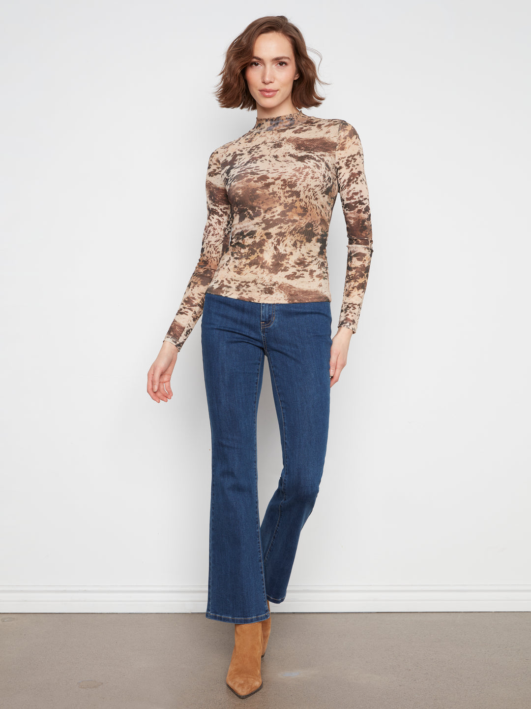 A woman modeling a long-sleeve mock neck top with a brown and beige abstract camouflage pattern, paired with blue jeans and tan ankle boots, standing against a white wall.