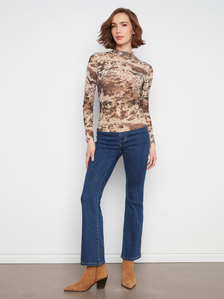A woman modeling a long-sleeve mock-neck top with a brown and beige abstract camouflage pattern, paired with blue jeans and tan ankle boots, standing against a white wall.