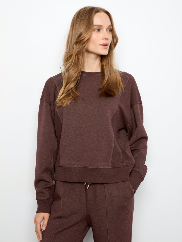 A woman wearing a dark brown crewneck sweatshirt and matching brown pants, photographed against a plain white studio background.