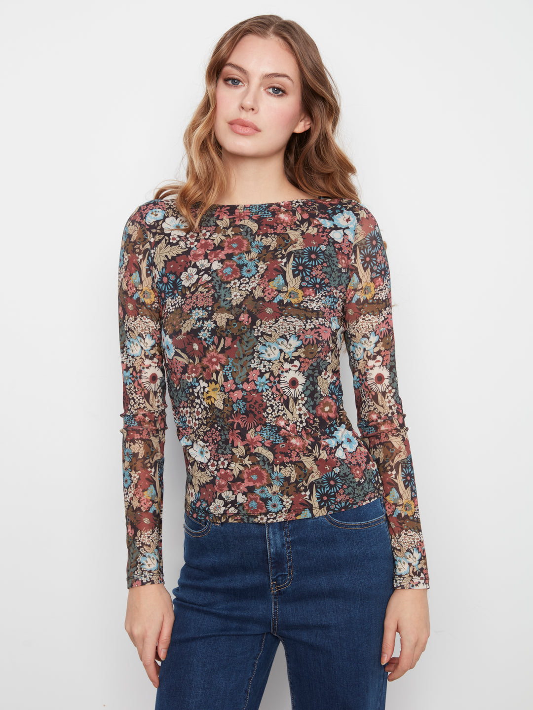 A woman modeling a long-sleeve boat-neck top with an all-over dense floral and botanical print in muted red, blue, green, beige, and brown tones, paired with dark blue jeans against a plain white studio background.
