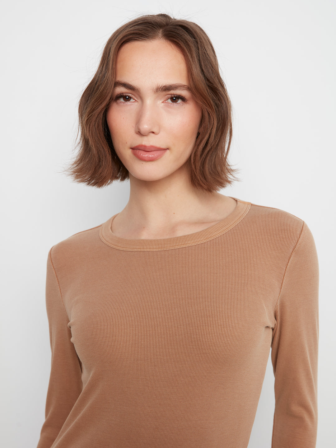 A woman with a shoulder-length brown bob haircut wearing a ribbed, long-sleeve crewneck top in a warm camel tan color, photographed against a plain light gray studio background.