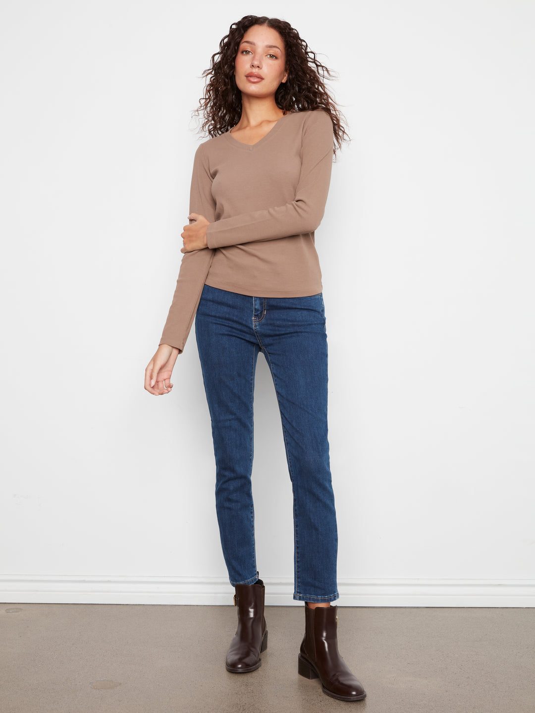 A woman modeling a beige long-sleeve V-neck knit top with dark blue skinny jeans and dark brown ankle boots, standing against a plain white wall.