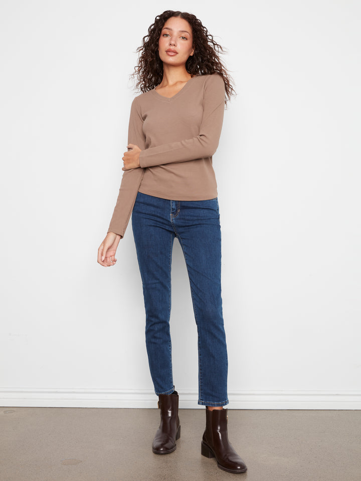 A woman modeling a beige long-sleeve V-neck knit top with dark blue skinny jeans and dark brown ankle boots, standing against a plain white wall.