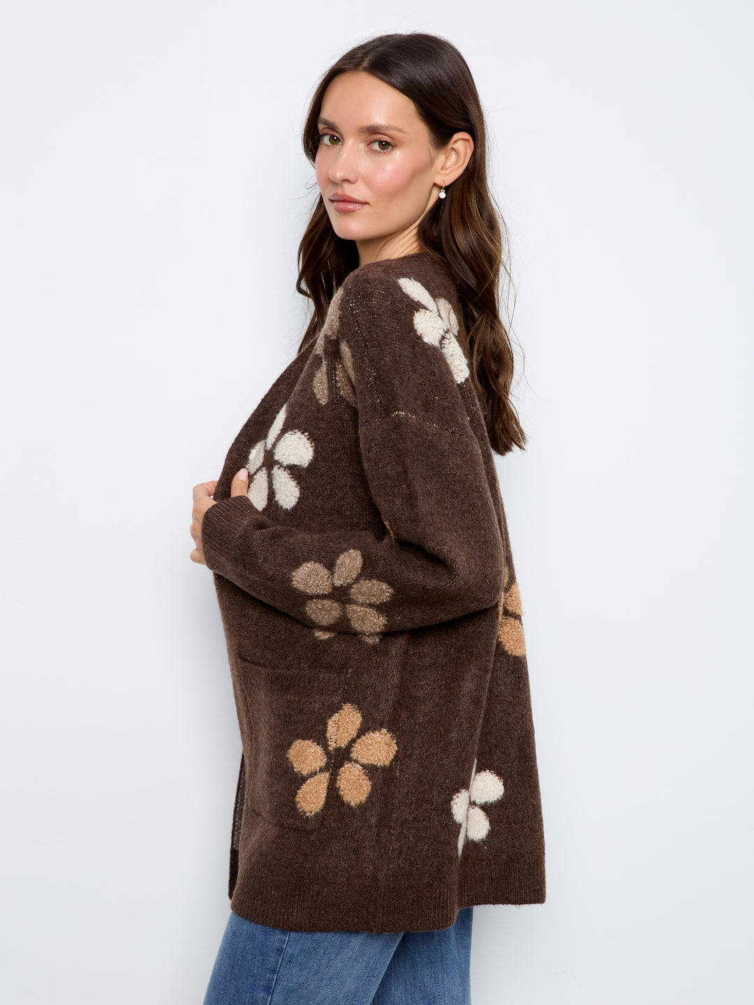 A woman wearing a brown fuzzy knit cardigan with large beige and white floral patterns, paired with blue jeans, photographed against a plain white studio background.