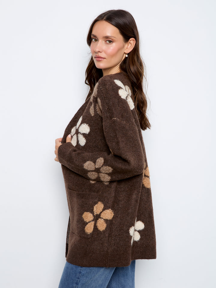 A woman wearing a brown fuzzy knit cardigan with large beige and white floral patterns, paired with blue jeans, photographed against a plain white studio background.