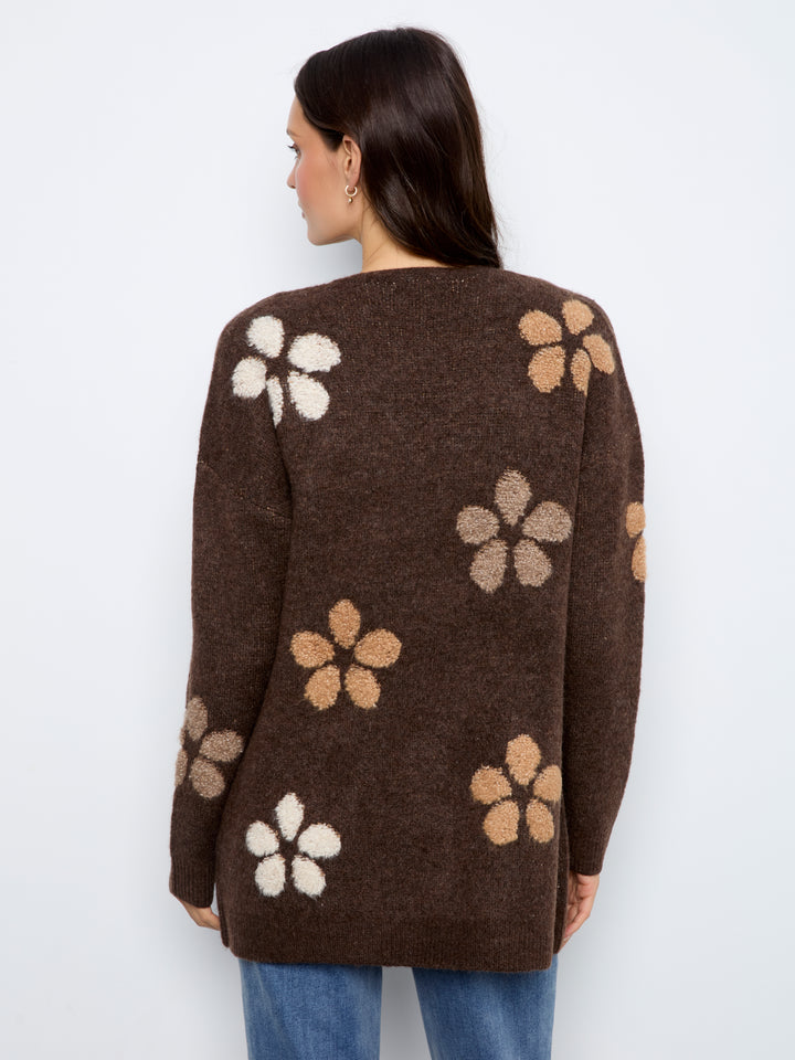 A woman viewed from the back wearing a brown knit sweater with large floral motifs in cream, tan, and light beige, paired with blue jeans against a plain light gray background.