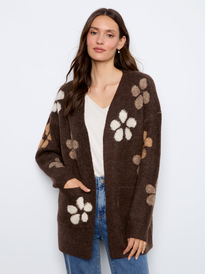 A woman modeling a brown open-front cardigan with large cream and beige flower motifs, worn over a white top and blue jeans against a light gray studio background.