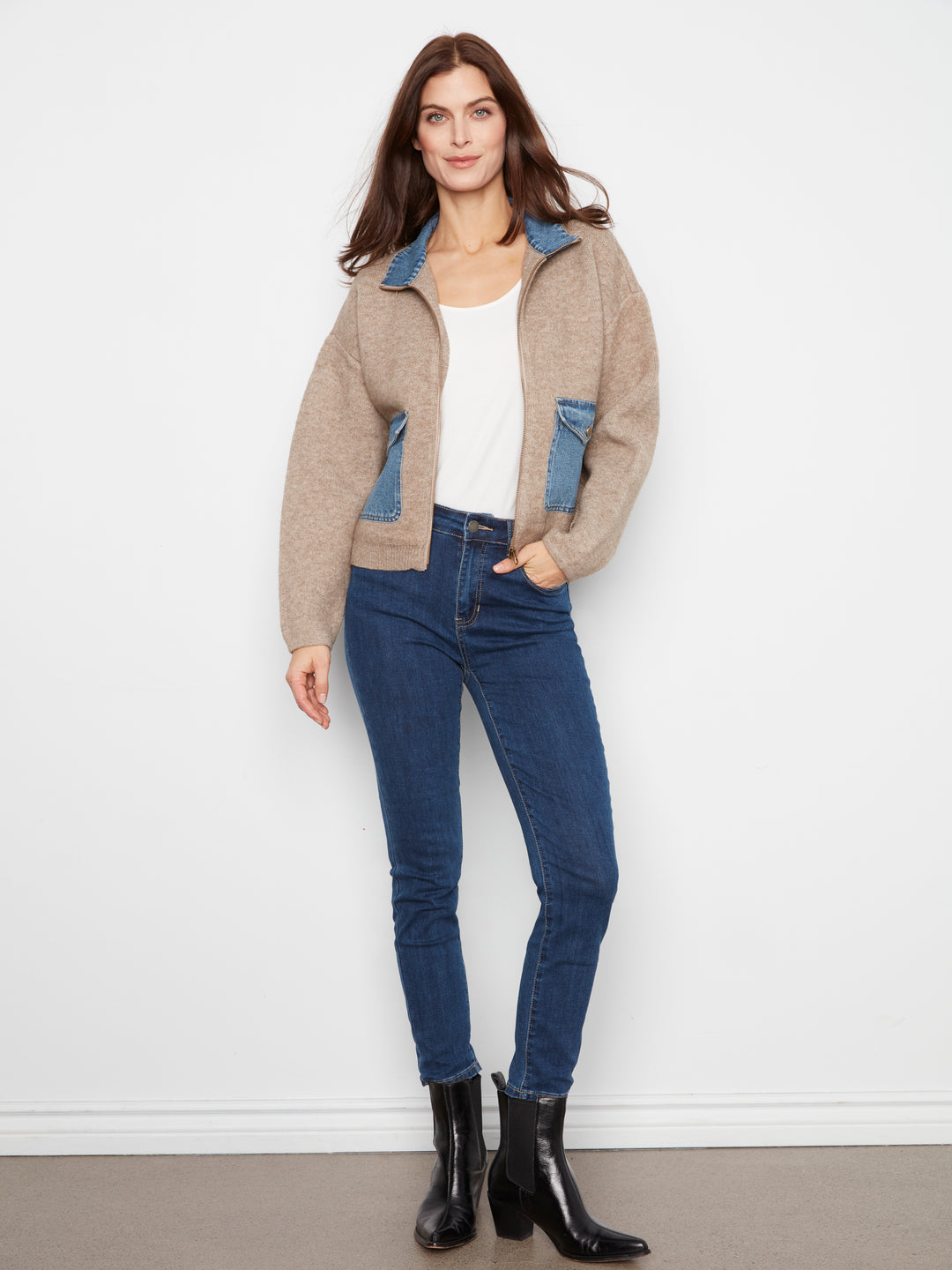 A woman modeling a beige knit zip-up hoodie jacket with blue denim pocket panels, worn over a white top and blue skinny jeans with black ankle boots, against a plain white wall.