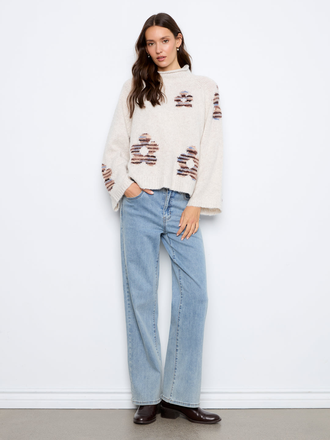 A woman stands against a white wall wearing a cream knit sweater with brown and black patterned accents, light blue wide-leg jeans, and dark brown shoes.