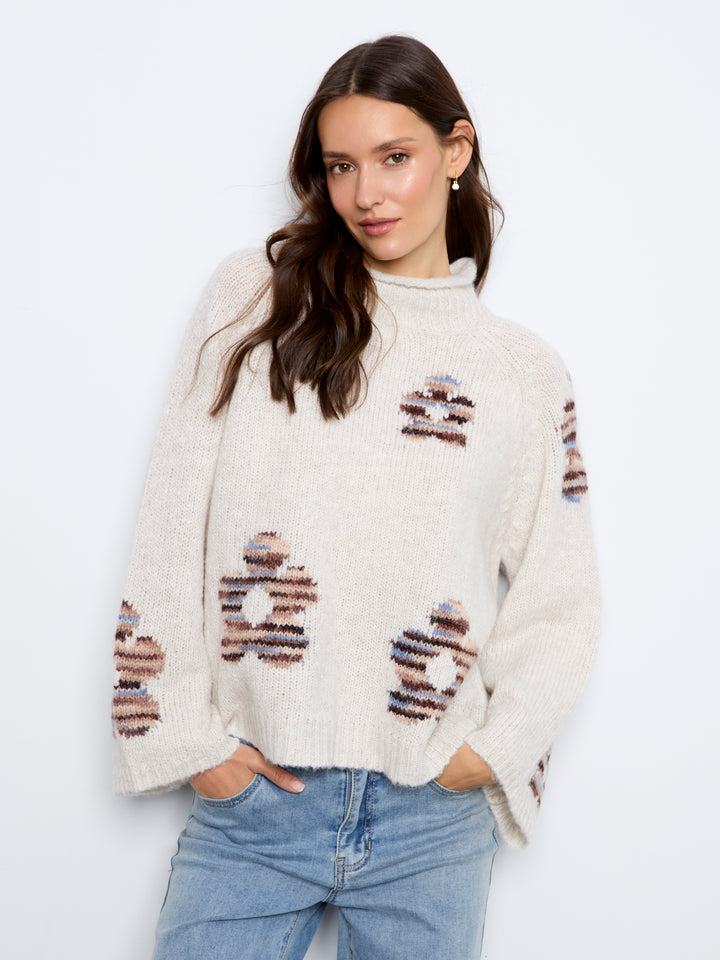 A woman wearing a cream-colored knit sweater with distressed patches of brown, blue, and white stripes, paired with light blue jeans, against a plain white background.