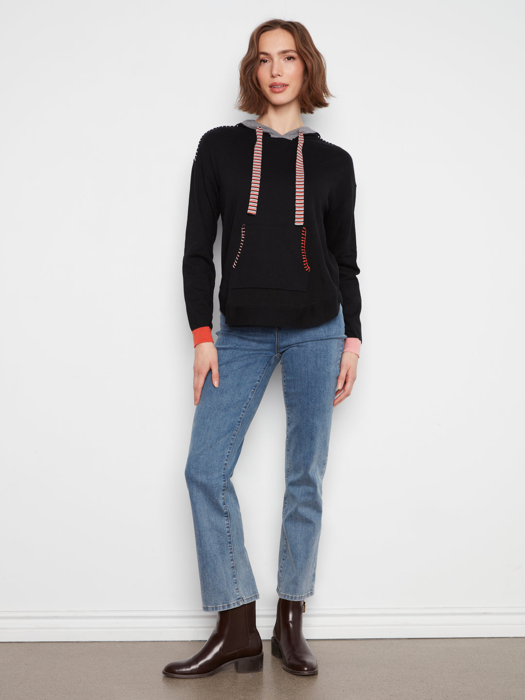 A woman modeling a black knit hoodie with red and white striped drawstrings, paired with blue jeans and dark brown ankle boots, standing against a plain white wall.