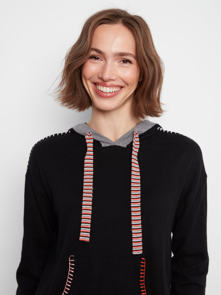 A smiling woman with short brown hair wearing a black knit hoodie with a gray hood lining and red-and-white striped drawstrings, photographed against a light gray studio background.