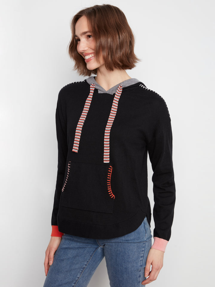 A woman wearing a black pullover hoodie with pink and white striped drawstrings, a gray hood lining, and pink ribbed cuffs, paired with blue jeans against a light gray background.