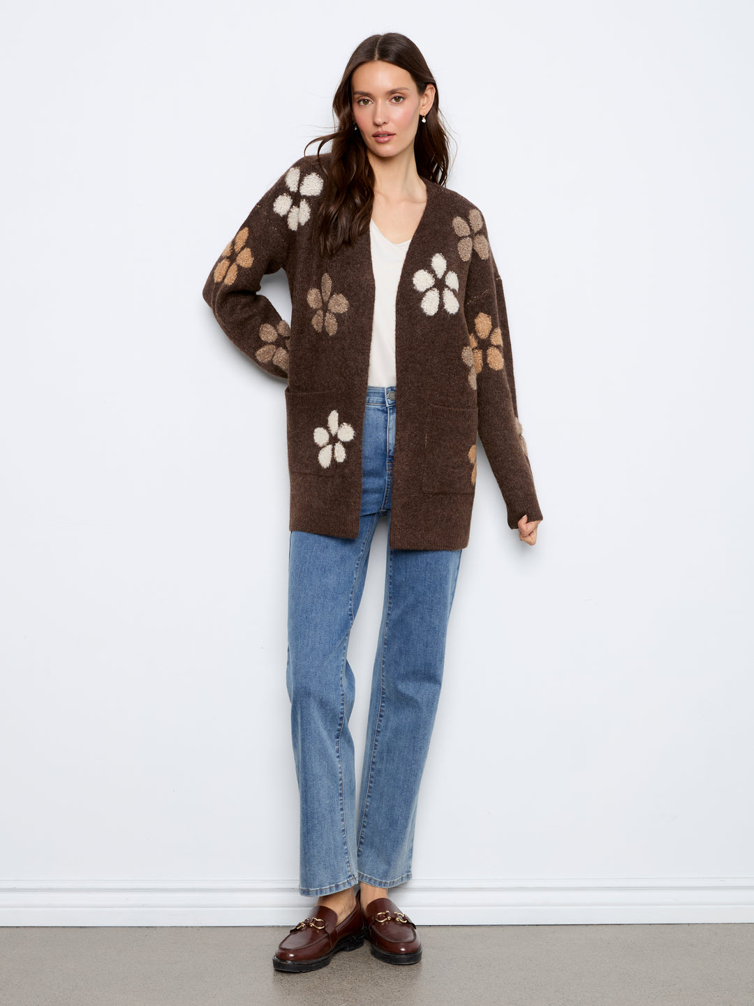A woman models a brown open-front cardigan with large white and tan floral patterns, paired with blue jeans, a white top, and brown loafers against a plain white wall.