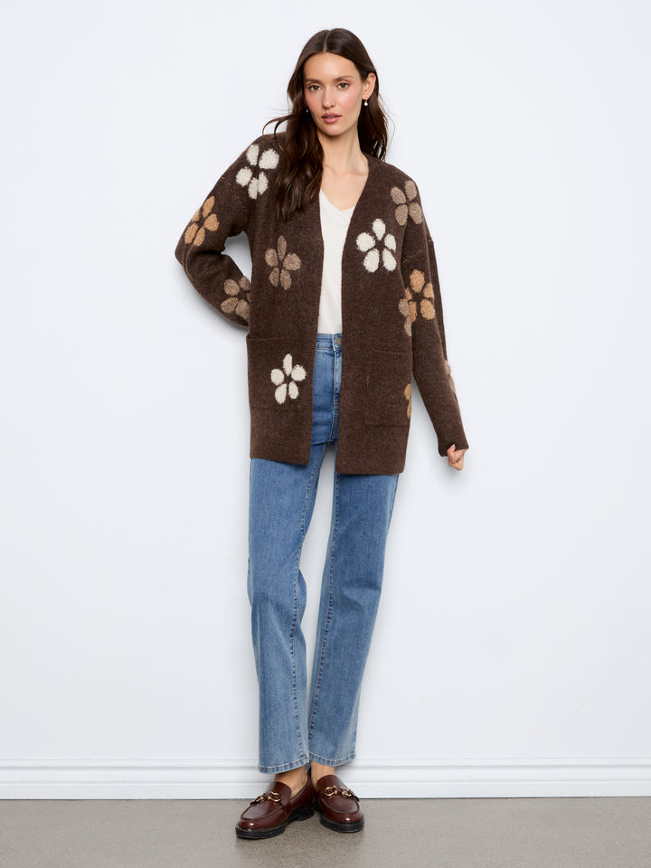 A woman models a brown open-front cardigan with large white and tan floral patterns, paired with blue jeans, a white top, and brown loafers against a plain white wall.