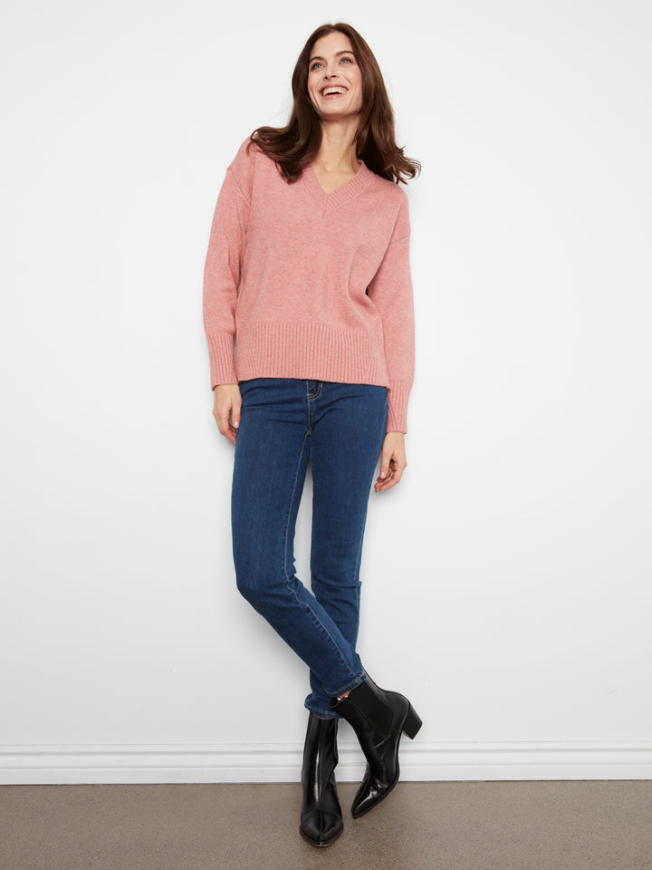 Woman wearing a pink sweater and blue jeans standing against a white wall.