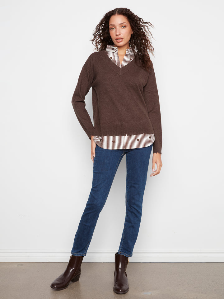 A woman modeling a brown v-neck knit sweater layered over a beige and white striped shirt, paired with blue jeans and dark brown ankle boots, against a plain white wall.