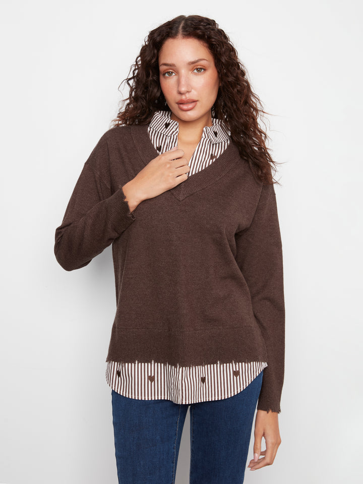 A woman modeling a brown knit sweater layered over a striped button-up shirt with blue jeans, photographed against a plain light gray studio background.