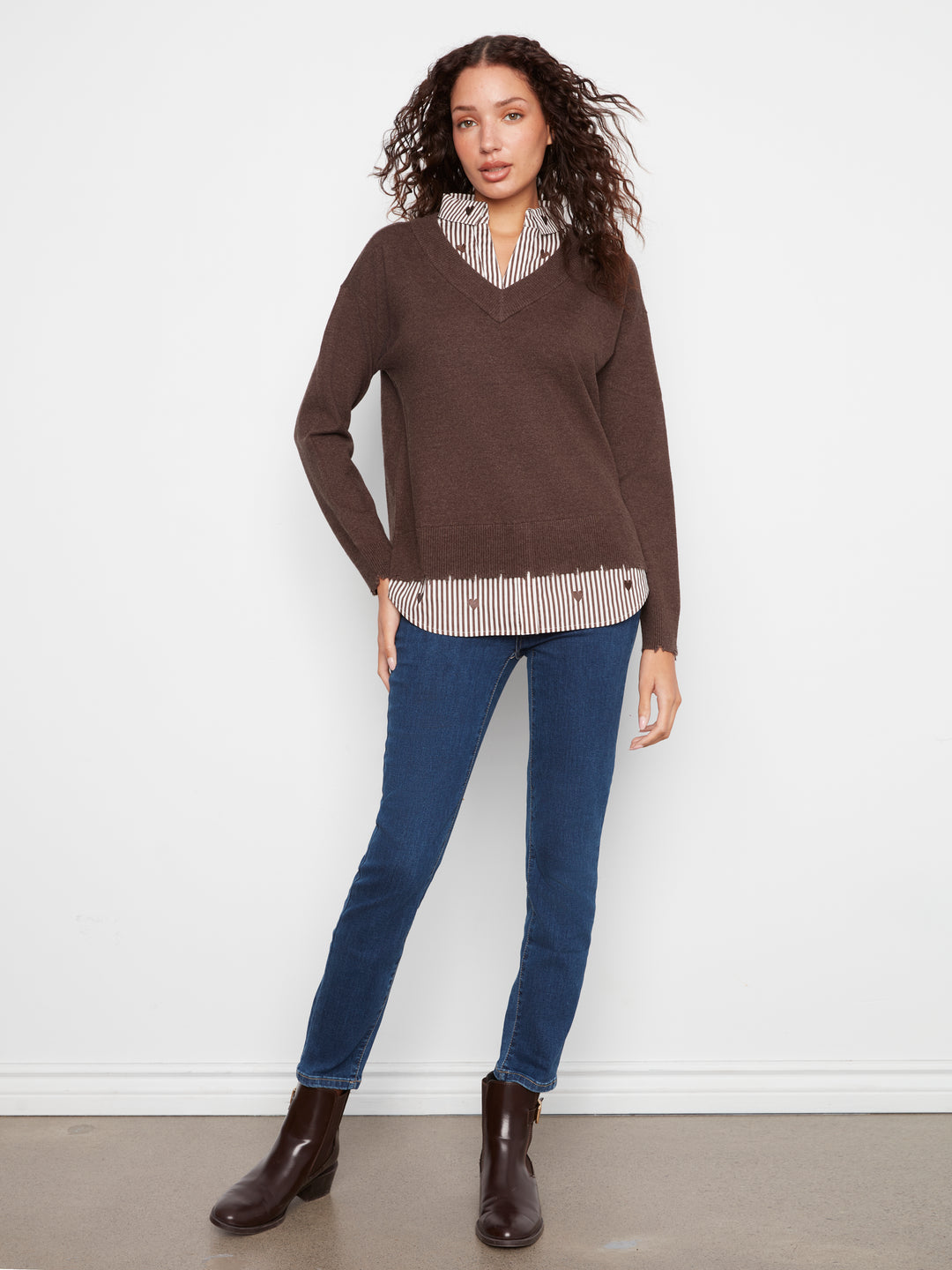 A woman modeling a brown V-neck knit sweater layered over a beige and white striped button-down shirt, paired with blue jeans and dark brown ankle boots, against a plain white wall.