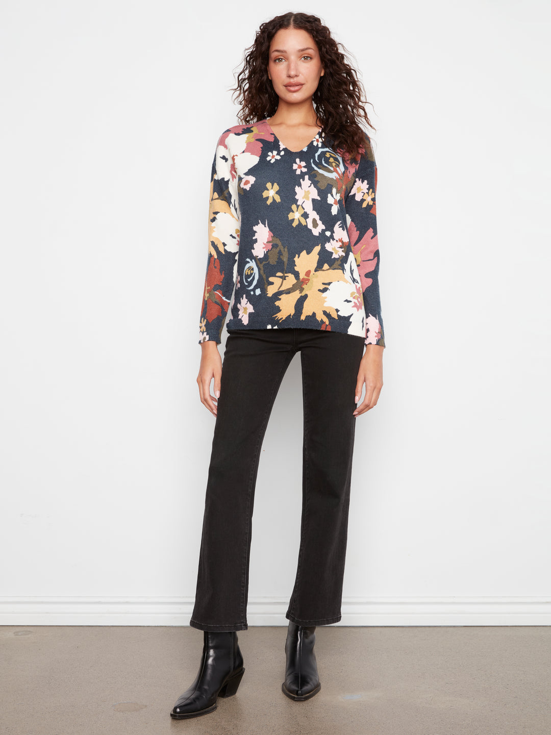 A woman stands against a white wall wearing a long-sleeve floral blouse with a dark navy base and multicolored floral print, paired with black pants and black ankle boots.