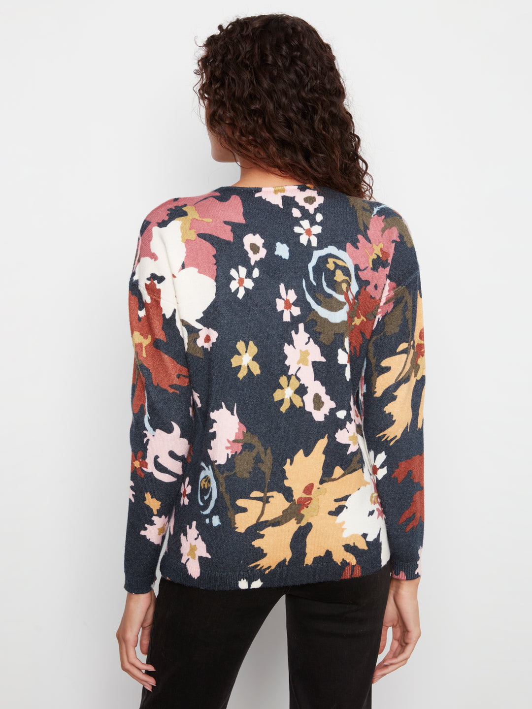 A woman viewed from the back wearing a long-sleeve knit top with a dark navy base and a large multicolor floral print in beige, pink, yellow, and brown, paired with black pants against a light gray studio background.