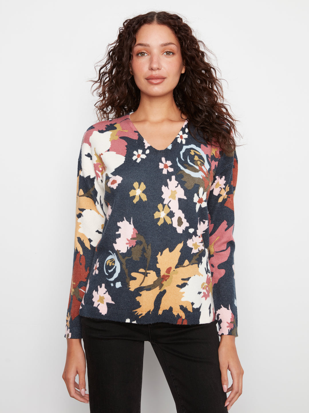A woman with curly brown hair wearing a long-sleeve V-neck top with a multicolor floral pattern on a dark navy base, paired with black pants, against a plain light gray background.