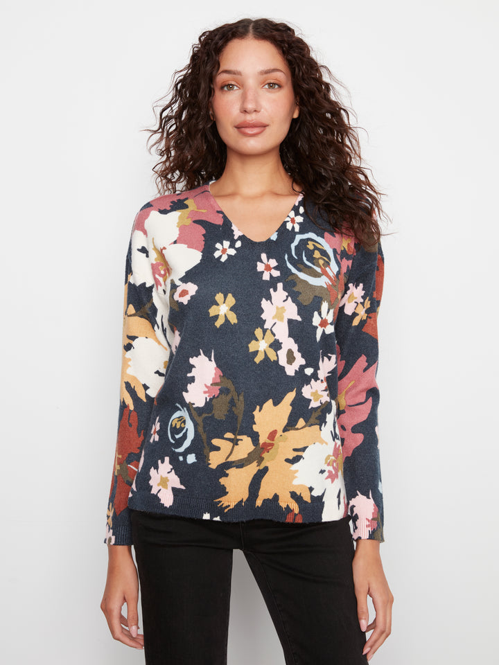 A woman with curly brown hair wearing a long-sleeve V-neck top with a multicolor floral pattern on a dark navy base, paired with black pants, against a plain light gray background.