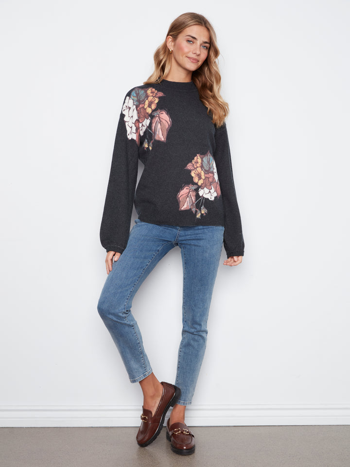 A woman modeling a black crewneck sweater with a large floral print on the front and shoulders, paired with blue jeans and brown loafers, standing against a white wall.