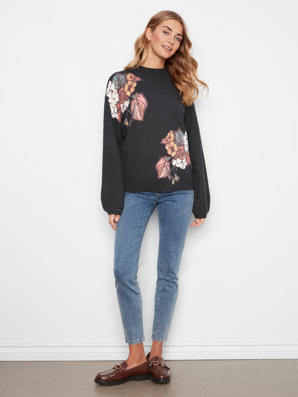 A woman modeling a black long-sleeve crewneck sweater with a large multicolor floral print on the front and one shoulder, paired with blue jeans and brown loafers, standing against a white wall.