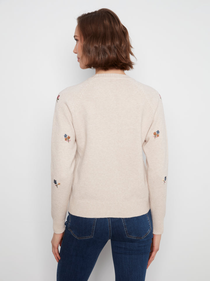 A woman viewed from the back wearing a cream-colored knit sweater with small embroidered floral details on the sleeves, paired with dark blue jeans, against a plain light gray background.