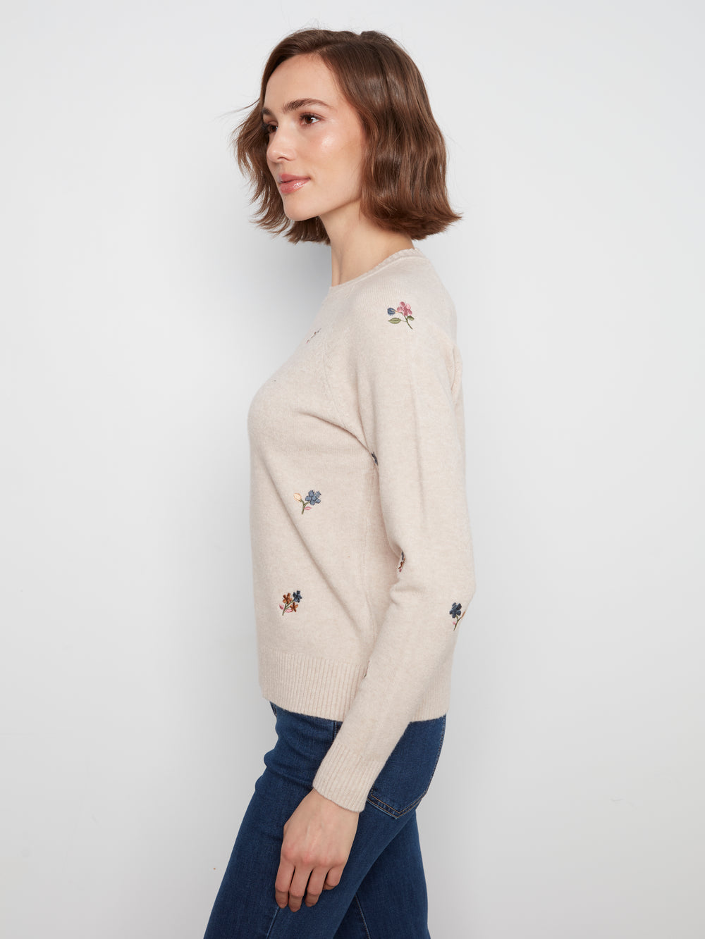 A woman wearing a light beige knit sweater with small embroidered floral details on the chest and sleeves, paired with dark blue jeans, shown from a side profile against a plain light gray background.