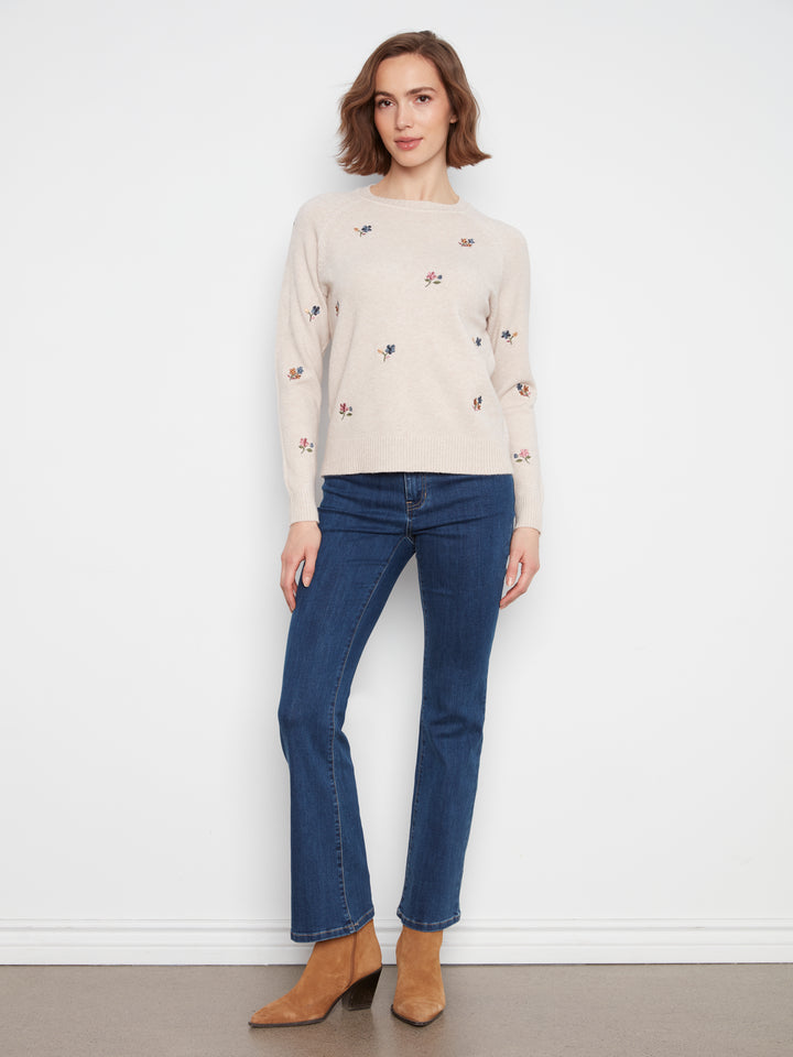 A woman stands against a plain white wall wearing a light beige long-sleeve knit sweater with small embroidered motifs, dark blue jeans, and tan ankle boots.