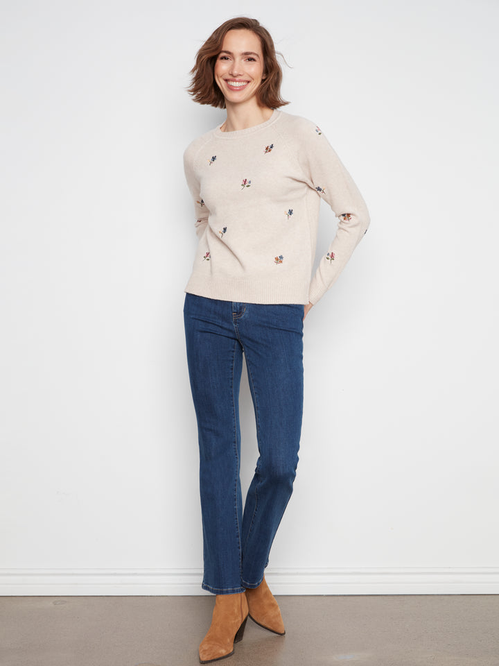 A woman modeling a light beige crewneck sweater with small embroidered details, paired with dark blue jeans and tan ankle boots, standing against a plain white wall.