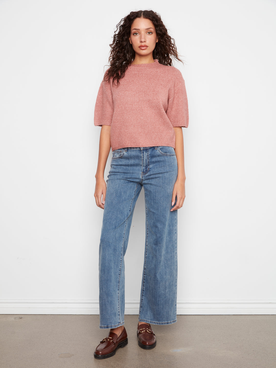 A woman stands against a plain white wall wearing a dusty rose short-sleeve knit top, blue wide-leg jeans, and brown leather loafers.