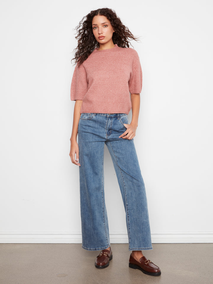 A woman modeling a dusty rose short-sleeve knit top and wide-leg blue jeans with brown leather loafers, standing against a plain white wall.