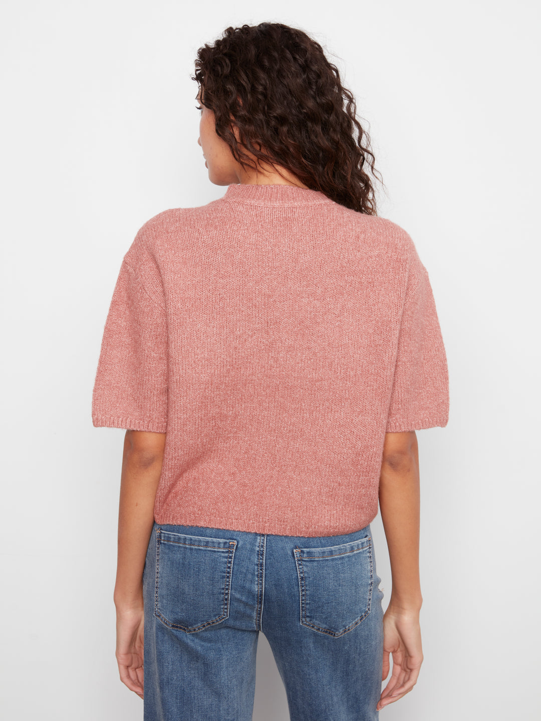 A woman viewed from the back wearing a dusty rose short-sleeve knit top and blue jeans against a plain white background.