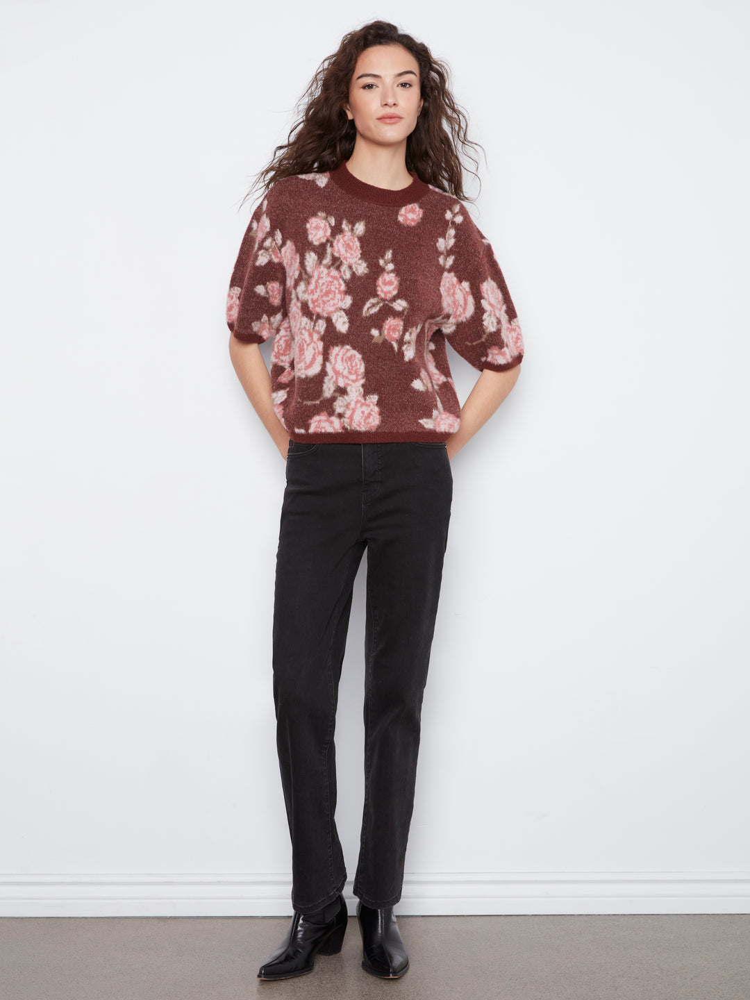 A woman modeling a short-sleeve crewneck sweater with a pink and beige floral pattern on a dark brownish-red background, paired with black trousers and black leather shoes, standing against a white wall.