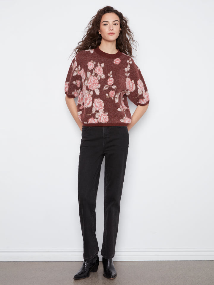 A woman modeling a short-sleeve crewneck sweater with a pink and beige floral pattern on a dark brownish-red background, paired with black trousers and black leather shoes, standing against a white wall.