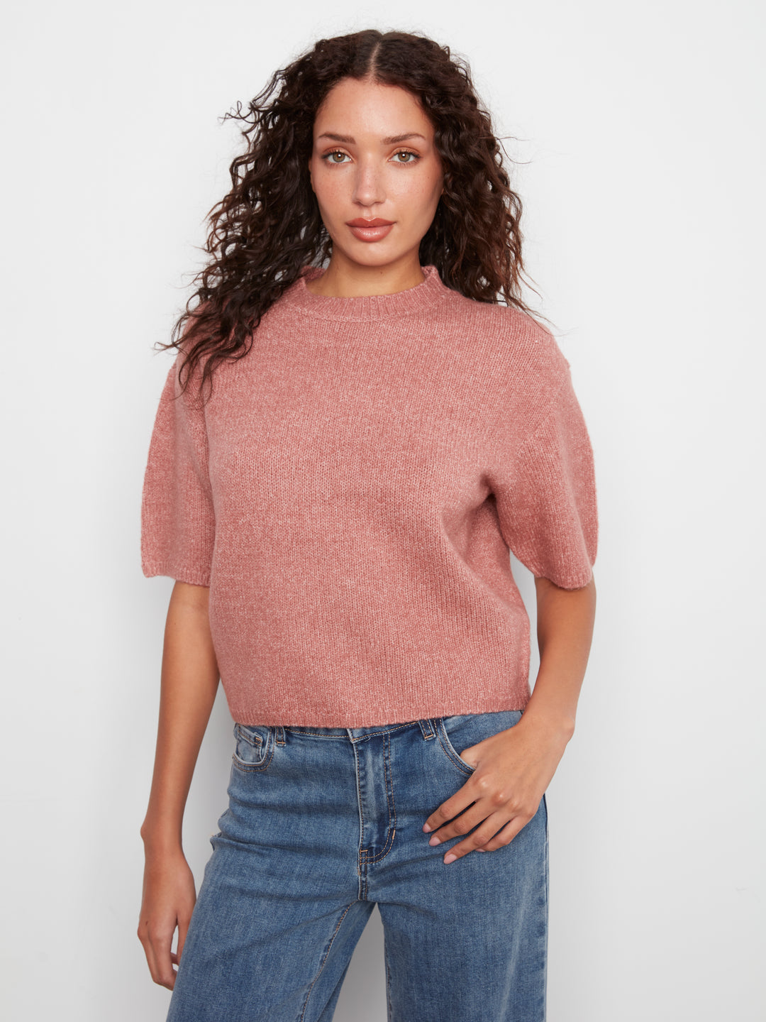 A woman with curly dark hair wearing a dusty rose short-sleeve knit top and blue jeans, standing against a plain light gray studio background.
