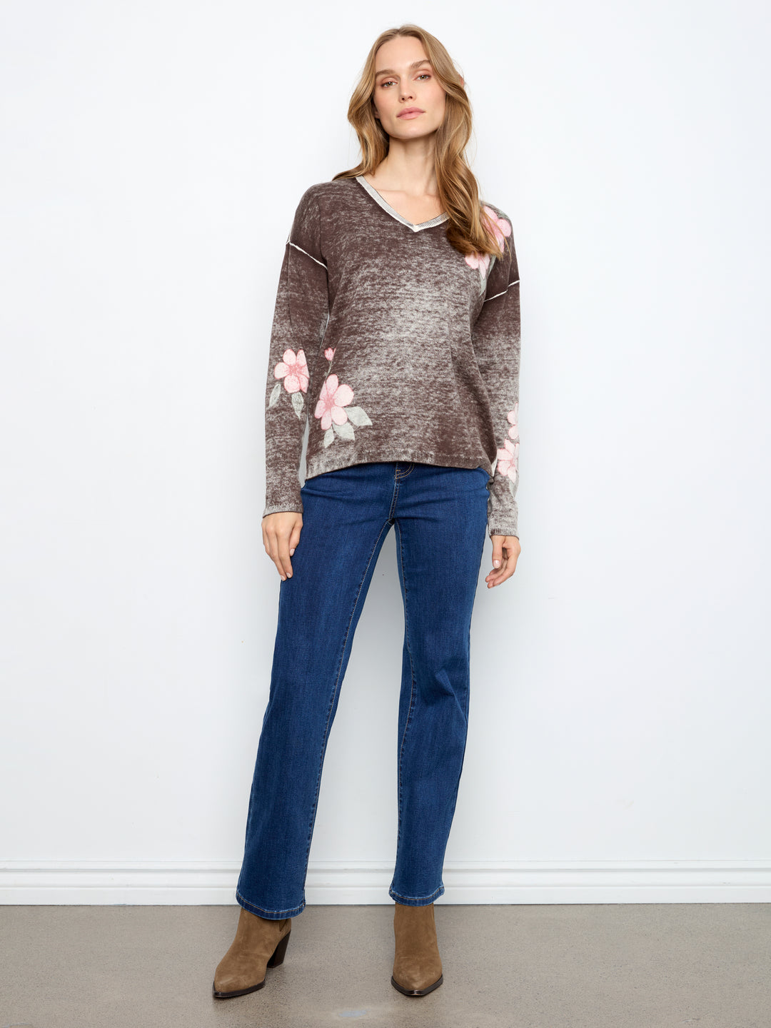 A woman modeling a brown and gray marled knit sweater with pink floral patterns on the sleeves and shoulders, paired with blue jeans and brown ankle boots, standing against a white wall.