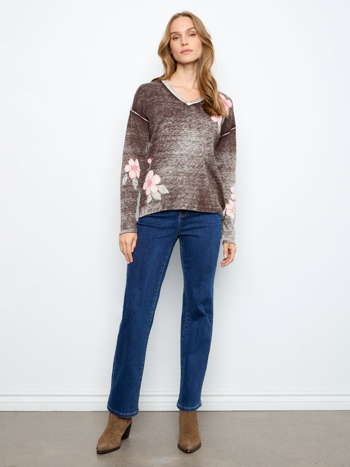 A woman modeling a brown and gray marled knit sweater with pink floral patterns on the sleeves and shoulders, paired with blue jeans and brown ankle boots, standing against a white wall.