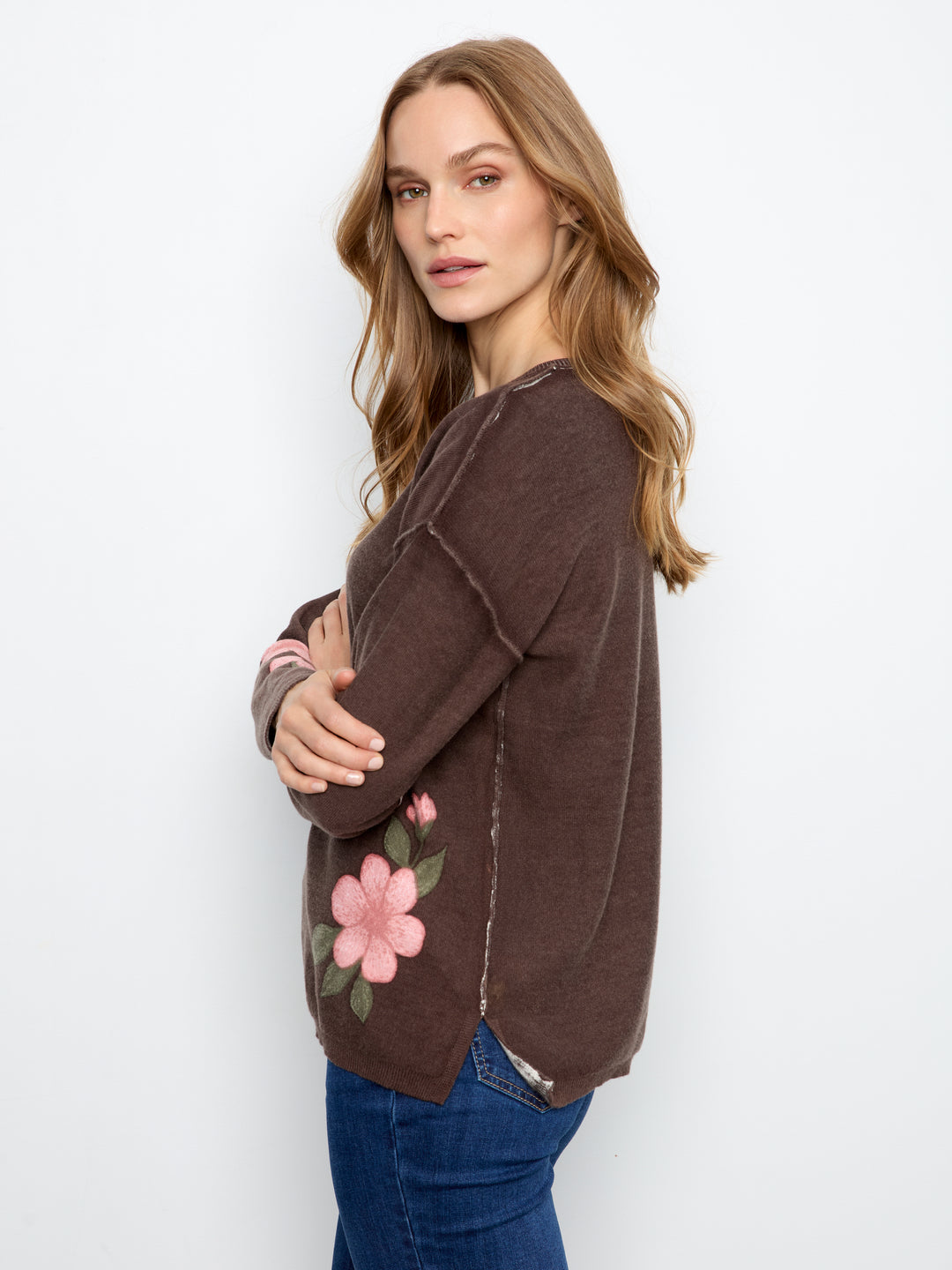 A woman wearing a brown long-sleeve knit sweater with a pink and green floral patchwork detail on the lower front, paired with blue jeans, photographed against a plain white studio background.