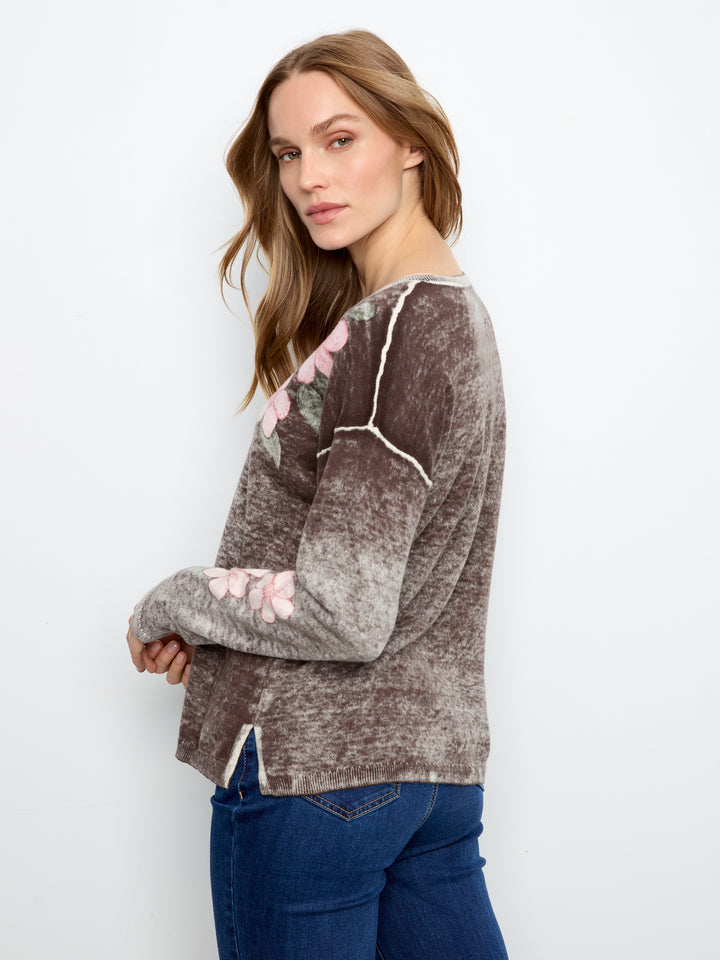 A woman wearing a brown and gray marled knit sweater with pink floral embroidery on the sleeves, paired with blue jeans, photographed against a plain white studio background.